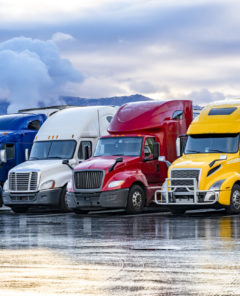 Different make big rigs semi trucks tractors with loaded semi trailers standing in the row on truck stop parking lot at early morning waiting for the route continuation time according to the log book