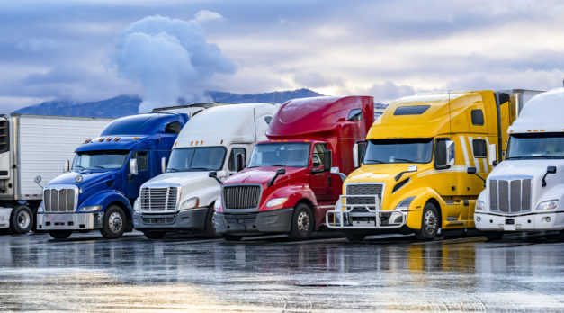 Different make big rigs semi trucks tractors with loaded semi trailers standing in the row on truck stop parking lot at early morning waiting for the route continuation time according to the log book