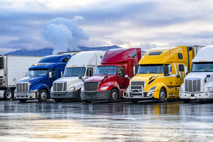 Different make big rigs semi trucks tractors with loaded semi trailers standing in the row on truck stop parking lot at early morning waiting for the route continuation time according to the log book